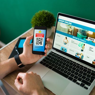 Young man using a discount coupon on his smartphone to do some online shopping on the laptop